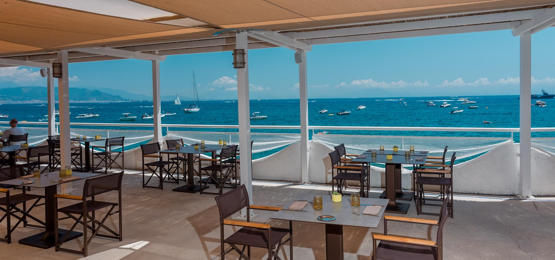 Ristorante con vista mare, tavoli all'aperto, barche in lontananza.